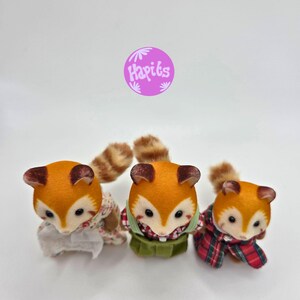 Puede incluir: Tres figuras de zorro con pelaje naranja y marr&oacute;n, cada una con un atuendo diferente. Una lleva un vestido floral, otra un chaleco verde y una bufanda a cuadros rojos, y la tercera una bufanda a cuadros rojos. Un c&iacute;rculo morado con el texto "Hapiks" est&aacute; en el fondo.