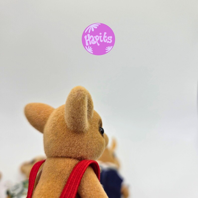 May include: A close-up of a tan-colored toy animal with a red strap. The toy has large ears and a small black eye. The word "Hapiks" is in a purple circle in the upper right corner.
