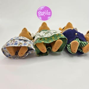 May include: A group of small, plush animal figures, viewed from the rear, wearing miniature clothing. One figure wears a blue floral skirt, another a green skirt with white lace, and a third a blue jacket with green striped pants. The figures are on a white background.