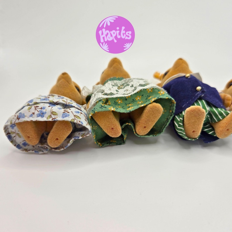 May include: A group of small, plush animal figures, viewed from the rear, wearing miniature clothing. One figure wears a blue floral skirt, another a green skirt with white lace, and a third a blue jacket with green striped pants. The figures are on a white background.