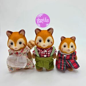 Puede incluir: Tres figuras de zorro de juguete con pelaje naranja y blanco, cada una con diferentes atuendos. Una lleva un vestido floral, otra una camisa a cuadros roja y un peto verde, y la tercera un vestido a cuadros rojo. Las figuras tienen ojos negros.