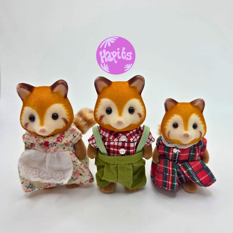 Puede incluir: Tres figuras de zorro de juguete con pelaje naranja y blanco, cada una con diferentes atuendos. Una lleva un vestido floral, otra una camisa a cuadros roja y un peto verde, y la tercera un vestido a cuadros rojo. Las figuras tienen ojos negros.