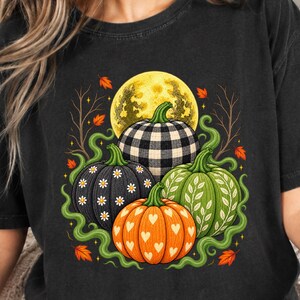 May include: A black short-sleeve t-shirt featuring a graphic print of four pumpkins beneath a yellow moon. The pumpkins display various patterns including black and white buffalo check, white daisies on black, white hearts on orange, and white leaves on green.