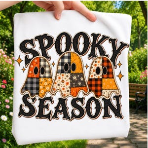 May include: A white t-shirt featuring a graphic of three ghosts with patchwork patterns in orange, black, and white. The text "SPOOKY SEASON" is printed in a bold, black, textured font above and below the ghosts, accented by small orange stars.