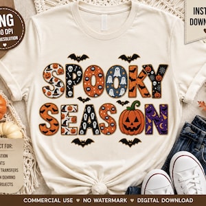 May include: A cream-coloured t-shirt featuring the text SPOOKY SEASON in large, patterned letters. The letters contain various Halloween motifs including pumpkins, ghosts, bats, skulls, and spiderwebs. Small black bat silhouettes surround the text.