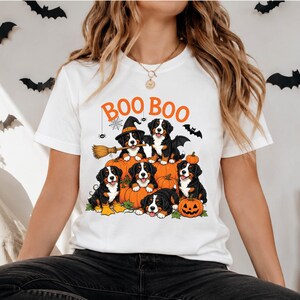 May include: A white t-shirt featuring a graphic print of six Bernese Mountain Dog puppies sitting among orange pumpkins. The text "BOO BOO" is printed in orange above the puppies. One puppy wears a witch hat and holds a broomstick.