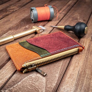May include: A multi-colored leather wallet with a zipper closure. The wallet is made of different shades of brown, green, and red leather. The zipper is gold-colored and has a pull tab. The wallet is lying on a wooden surface.