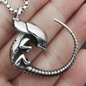 Könnte beinhalten: Eine silberfarbene Halskette mit Anhänger, die ein detailliertes Alien-Design zeigt. Der Anhänger zeigt einen Xenomorph mit gebogenem Schwanz und einer Kette. Das Alien befindet sich in einer hockenden Position mit offenem Mund. Die Halskette ist aus glänzendem Metall.