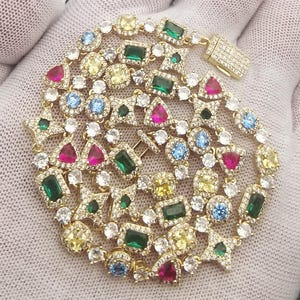 May include: A gold-toned bracelet with a variety of colorful gemstones, including green, red, blue, and yellow stones. The bracelet features a mix of shapes, including squares, triangles, and round stones, and a secure clasp.