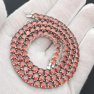 May include: A silver necklace with numerous round, red gemstones. The necklace is displayed on a white surface, showcasing the vibrant color and intricate design. The clasp is visible, highlighting the necklace's secure closure.
