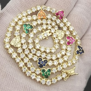 May include: A gold-toned necklace with a series of clear, round-cut stones. Interspersed are heart-shaped pendants in various colors, including green, blue, pink, and orange. The necklace is displayed on a white surface.