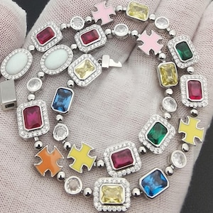 Può includere: Un braccialetto d'argento con una varietà di pietre preziose colorate di forma rettangolare, ovale e a croce. Le pietre sono incastonate in una fascia d'argento e circondate da piccole pietre trasparenti. Il braccialetto è esposto su una superficie bianca.