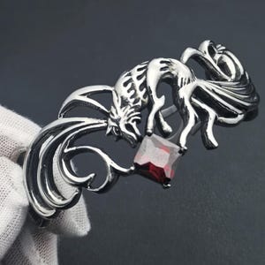 May include: Silver-toned bracelet featuring a stylized fox design with a red square gemstone. The bracelet has intricate details and a polished finish. The fox is depicted in a dynamic pose, adding a touch of fantasy.