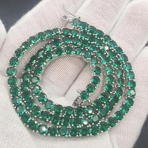 Könnte beinhalten: Eine silberfarbene Tennis-Halskette mit einer durchgehenden Reihe von rund geschliffenen, leuchtend grünen Steinen in einer Metallfassung. Die Kette ist kreisförmig gewickelt und zeigt die einheitliche Größe und leuchtend grüne Farbe der Steine.