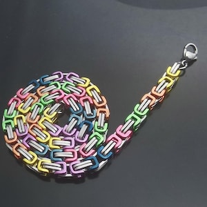 Op de afbeelding: Een veelkleurige ketting met een roestvrijstalen sluiting. De ketting bestaat uit in elkaar grijpende rechthoekige schakels in een regenboog van kleuren, waaronder roze, geel, groen en blauw. De ketting wordt gepresenteerd op een donkere, reflecterende ondergrond.