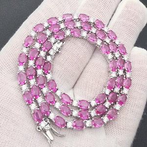 Puede incluir: Un collar de plata con piedras preciosas rosas ovaladas y pequeñas piedras blancas. Las piedras están engastadas muy juntas, creando una línea continua de color. El collar se muestra sobre una superficie blanca.