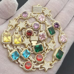 May include: Gold-toned necklace with a circular design, featuring a variety of colorful gemstones in square and round shapes. The necklace is adorned with small star-shaped accents and clear crystals, creating a sparkling effect.
