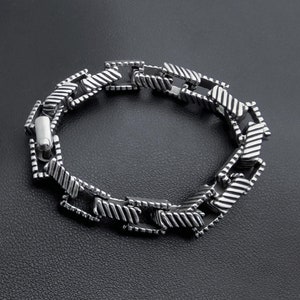 Könnte beinhalten: Ein silbernes Kettenarmband mit einem strukturierten, gemusterten Design. Das Armband besteht aus Metallgliedern, die miteinander verbunden sind, um eine Kette zu bilden.