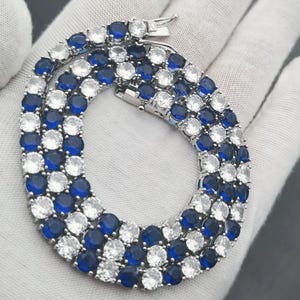 May include: A tennis necklace featuring a repeating pattern of round, brilliant-cut clear white stones and deep blue stones set in a silver-toned metal frame. The piece includes a secure box clasp closure and a flexible, articulated chain design.