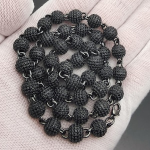 Puede incluir: Collar de cuentas negro con un diseño esférico texturizado. El collar está hecho de pequeñas cuentas oscuras unidas por una cadena y un cierre a juego. La joya se muestra sobre una superficie blanca.