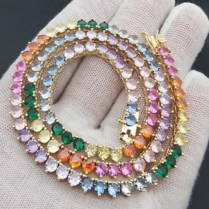 Pode incluir: Um colar dourado com uma variedade de pedras preciosas coloridas. As pedras são dispostas em um padrão circular, mostrando um gradiente de cores do rosa ao azul, verde e amarelo. O colar é exibido em uma superfície branca.
