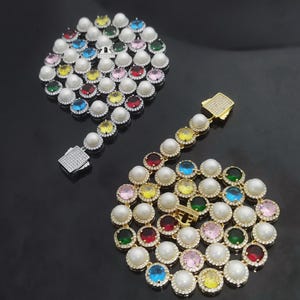 Op de afbeelding: Twee kettingen met afwisselend ronde witte parels en ronde veelkleurige edelstenen, waaronder rode, blauwe, gele, groene en roze stenen. Elke edelsteen is omringd door kleine heldere kristallen en de kettingen hebben een doosvormige sluiting.