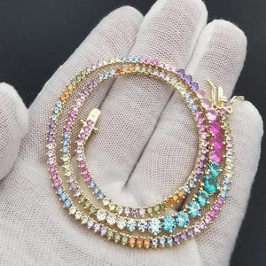 Puede incluir: Un collar dorado con múltiples hilos, cada uno adornado con una vibrante variedad de gemas de colores. Las gemas incluyen tonos de rosa, azul, naranja y verde, creando un efecto arcoíris. El collar se muestra sobre una superficie blanca.
