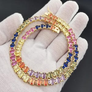 Puede incluir: Un collar dorado con gemas rectangulares multicolores. Las gemas presentan un arcoíris de colores, incluyendo azul, rosa, amarillo y naranja. El collar se muestra sobre una superficie blanca.