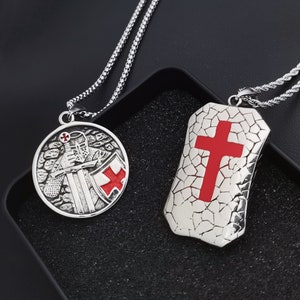 Op de afbeelding: Twee zilveren kettingen met hangers. De ene hanger heeft een ridder met een zwaard en schild, de andere een schild met een rood kruis. Beide hangers hebben een getextureerd oppervlak.