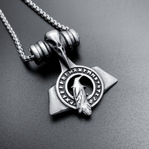 Puede incluir: Un collar de plata con un colgante que presenta un cuervo y un círculo de runas. El colgante tiene forma de martillo.