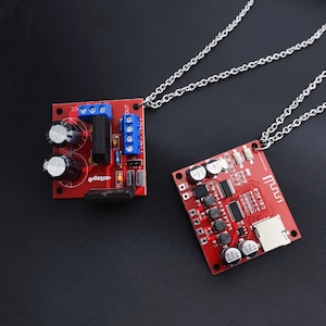 May include: Two red circuit boards with electronic components are attached to silver chains. The circuit boards are connected to a silver chain necklace. The circuit boards have text on them, including "Output" and "Music".