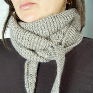 Può includere: Un primo piano di una sciarpa di maglia marrone chiaro indossata intorno al collo. La sciarpa è realizzata con un filato morbido e testurizzato ed è piegata su se stessa per creare un look a strati.