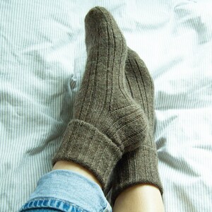 May include: A pair of brown ribbed socks with a folded cuff. The socks are worn on a person's feet and are resting on a white and gray striped bed sheet.