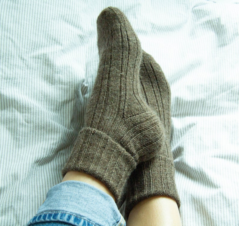 May include: A pair of brown ribbed socks with a folded cuff. The socks are worn on a person's feet and are resting on a white and gray striped bed sheet.