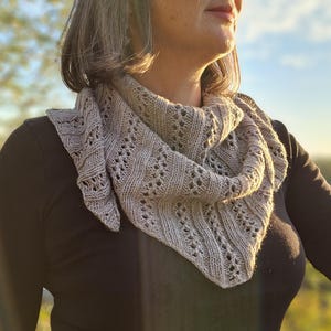 May include: A triangular knit scarf in a light beige color featuring a textured pattern of vertical ribbed columns alternating with delicate eyelet lace rows. The scarf is draped around the neck, showcasing the intricate yarn work and soft edges.
