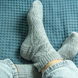 Könnte beinhalten: Ein Paar graue Stricksocken mit einem strukturierten Muster. Die Socken werden an den Füßen einer Person getragen und sind teilweise auf einer blauen strukturierten Oberfläche sichtbar.