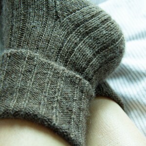 May include: A close-up of a brown knitted sock with a ribbed cuff. The sock is on a white and gray striped surface.