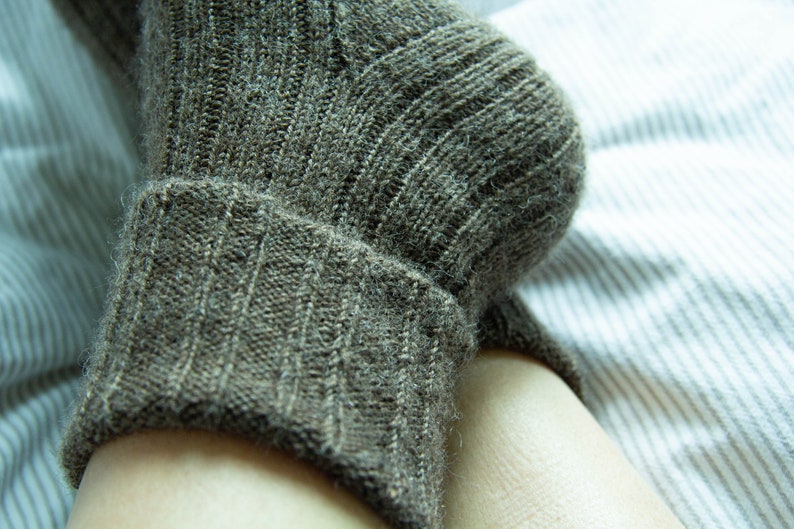 May include: A close-up of a brown knitted sock with a ribbed cuff. The sock is on a white and gray striped surface.