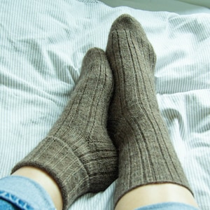 May include: A pair of brown wool socks with a ribbed texture, worn on a person's feet. The socks are on a white and gray striped bed sheet.