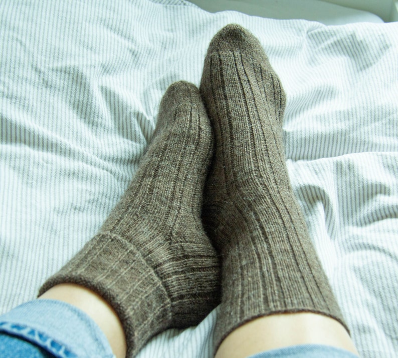 May include: A pair of brown wool socks with a ribbed texture, worn on a person's feet. The socks are on a white and gray striped bed sheet.