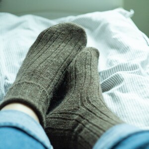 May include: A pair of brown wool socks with ribbed texture, worn on a person's feet, resting on a white and gray striped bed sheet.