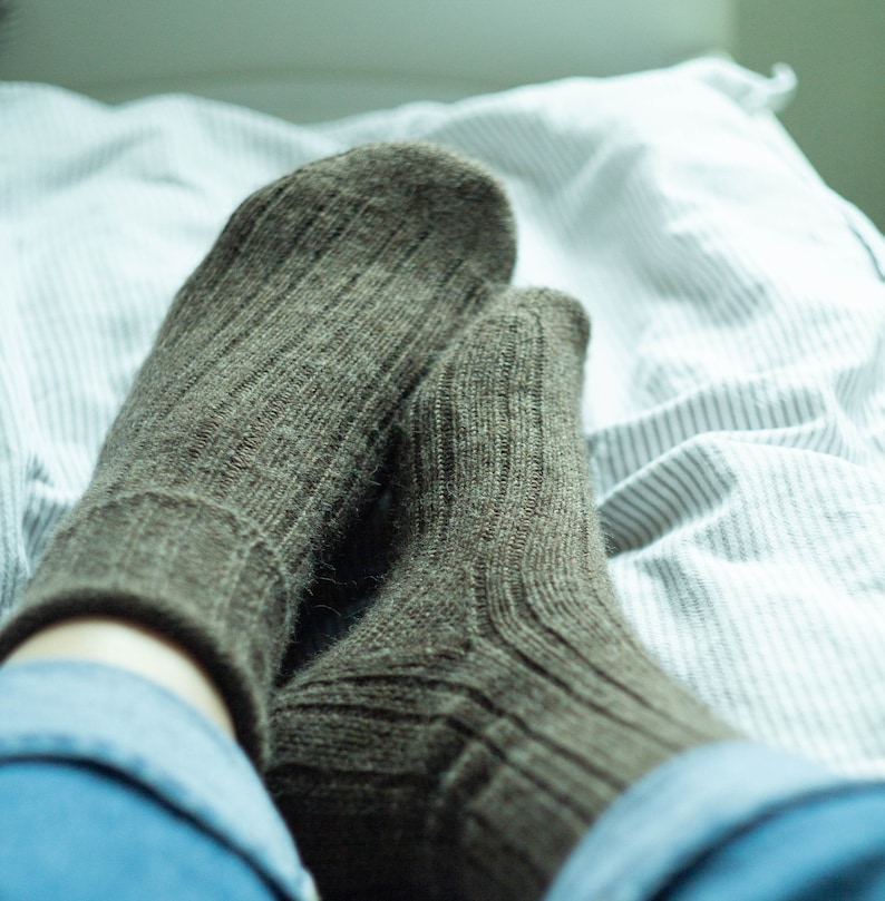 May include: A pair of brown wool socks with ribbed texture, worn on a person's feet, resting on a white and gray striped bed sheet.