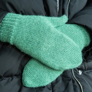 May include: A pair of green knitted mittens, folded and held in a person's hands. The mittens are made of a soft, fuzzy yarn.