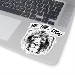 May include: A white sticker featuring a black and white lion illustration with the text "BE THE LION" above it. The sticker is placed on a silver laptop. The lion's face is detailed and the text is in a handwritten style.