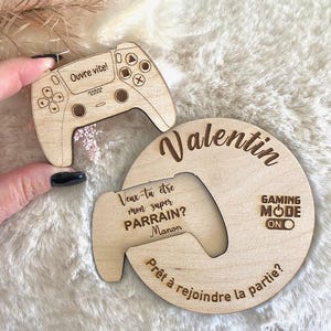 Könnte beinhalten: Dekorationen aus Holz zum Thema Gaming. Eines ist ein Controller mit dem Text "Ouvre vite!" und ein anderes ist ein kreisförmiges Stück mit dem Namen "Valentin" und dem Text "Gaming Mode On". Der Text "Veux-tu etre mon super PARRAIN? Manon" ist ebenfalls sichtbar.