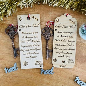 Könnte beinhalten: Zwei hölzerne Weihnachts-Schlüsselanhänger mit einem Schlüsselanhänger in Form eines Weihnachtsmanns. Die Anhänger sind mit dem Text "Cher Père Noël" und einer Botschaft über einen magischen Schlüssel graviert, der es dem Weihnachtsmann ermöglicht, das Haus zu betreten, um Geschenke zu liefern. Die Anhänger sind mit einer Schneeflocke und einem Herzen verziert.