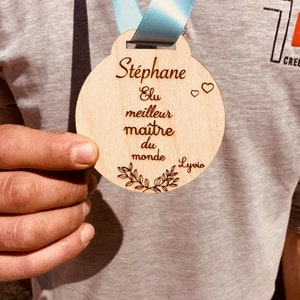 May include: A wooden medal with the text "Stephane Elu meilleur maitre du monde Lyvio" and two hearts. The medal is brown and has a light blue ribbon.
