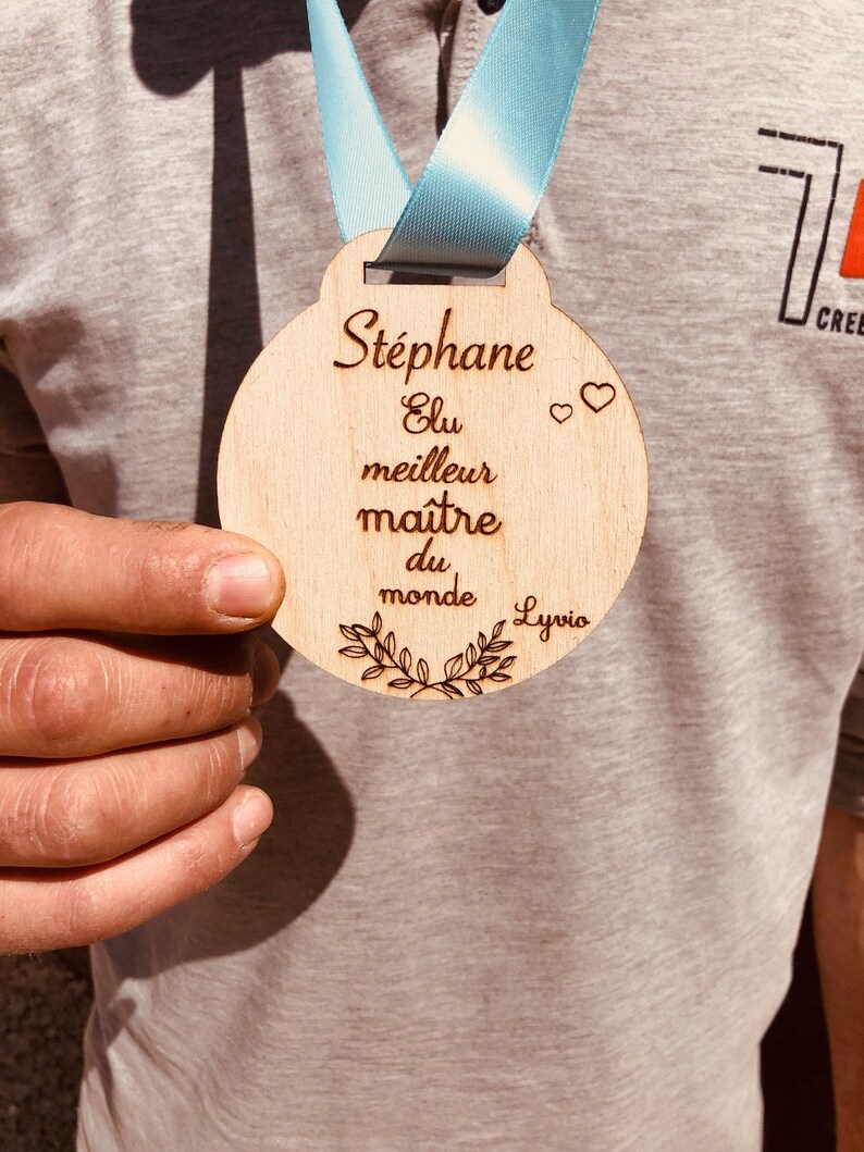 May include: A wooden medal with the text "Stephane Elu meilleur maitre du monde Lyvio" and two hearts. The medal is brown and has a light blue ribbon.
