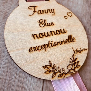 May include: A round wooden plaque with a pink ribbon attached. The plaque is engraved with the text "Fanny Elue nounou exceptionnelle kevin".
