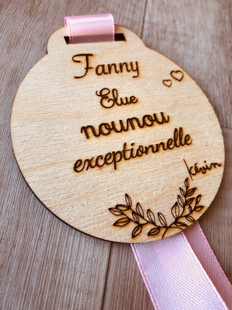 May include: A round wooden plaque with a pink ribbon attached. The plaque is engraved with the text "Fanny Elue nounou exceptionnelle kevin".